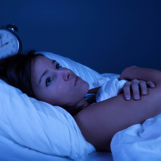 Woman struggling to fall asleep - has insomnia & is lying on white sweet sheets with an alarm clock